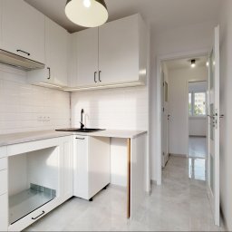 WYNAJMO SPÓŁKA Z OGRANICZONĄ ODPOWIEDZIALNOŚCIĄ - Jasna kuchnia z białymi szafkami i czarną armaturą we Wrocławiu, widok na korytarz z oknem w tle.