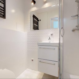 WYNAJMO SPÓŁKA Z OGRANICZONĄ ODPOWIEDZIALNOŚCIĄ - Jasna łazienka z kabiną prysznicową, umywalką z szafką i grzejnikiem drabinkowym, białe płytki na ścianach, minimalistyczny styl.