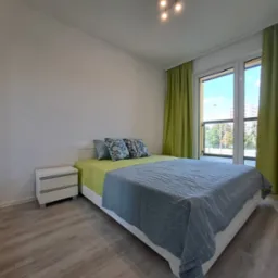 Jasna sypialnia z podwójnym łóżkiem, zielonymi zasłonami i szafką nocną, widok z perspektywy podłogi, z oknem wychodzącym na zieleń.