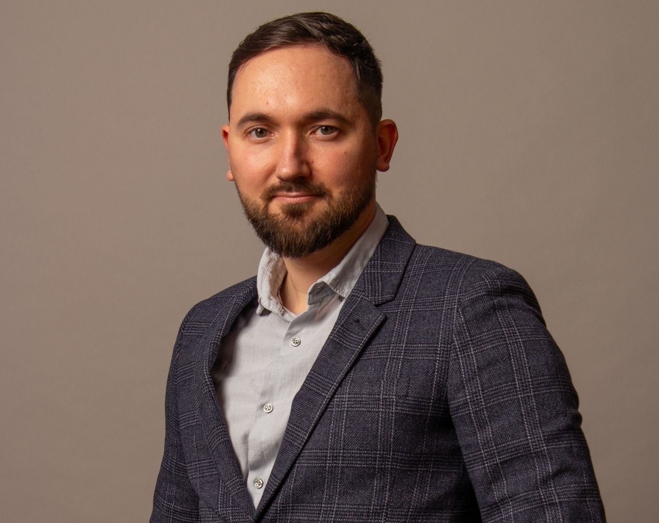 Portret mężczyzny z brodą w ciemnej marynarce w kratę i jasnej koszuli na jednolitym, szarym tle.