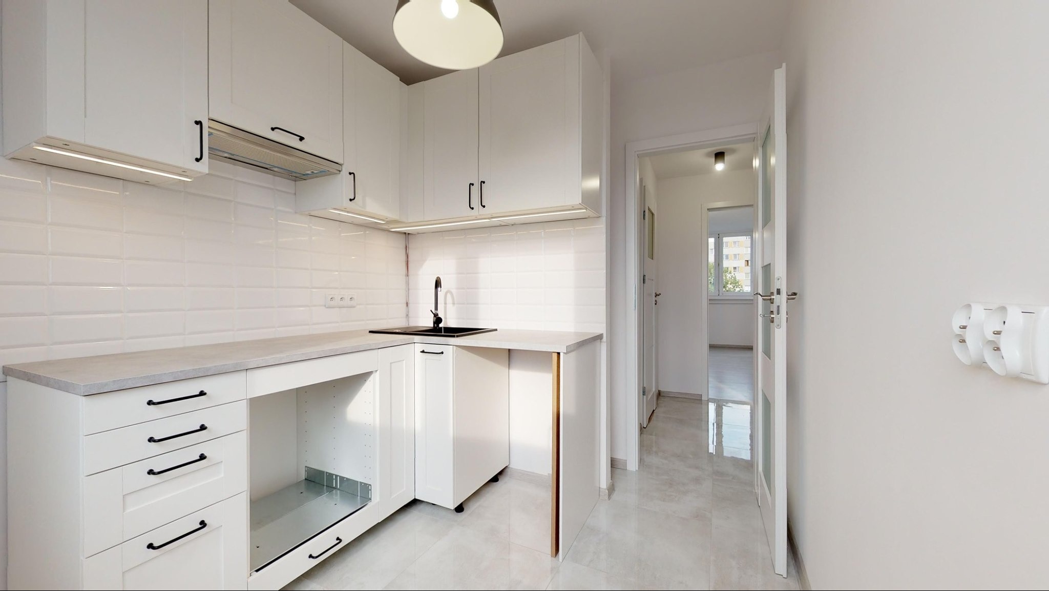 Jasna kuchnia z białymi szafkami i czarną armaturą we Wrocławiu, widok na korytarz z oknem w tle.