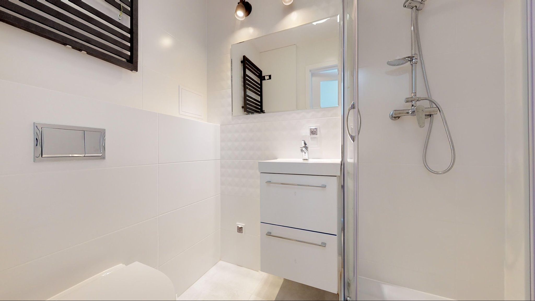 Jasna łazienka z kabiną prysznicową, umywalką z szafką i grzejnikiem drabinkowym, białe płytki na ścianach, minimalistyczny styl.