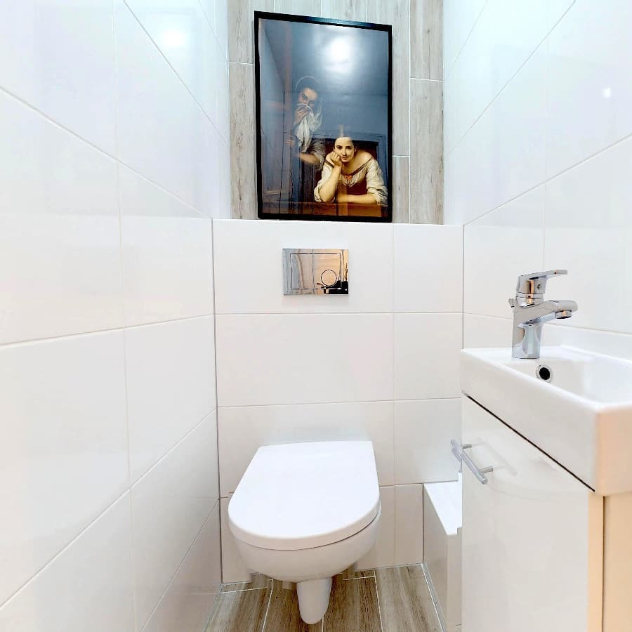 Nowoczesna, biała toaleta z podwieszaną muszlą, umywalką z szafką i chromowaną armaturą, ozdobiona obrazem w czarnej ramie, na tle białych płytek.