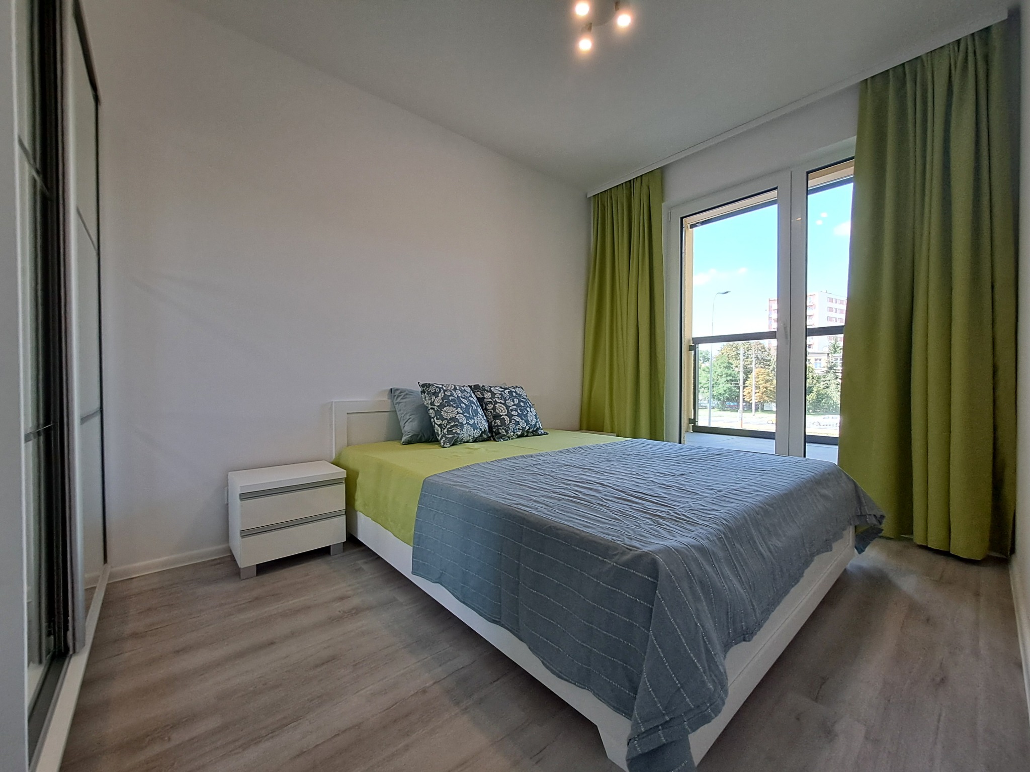 Jasna sypialnia z podwójnym łóżkiem, zielonymi zasłonami i szafką nocną, widok z perspektywy podłogi, z oknem wychodzącym na zieleń.