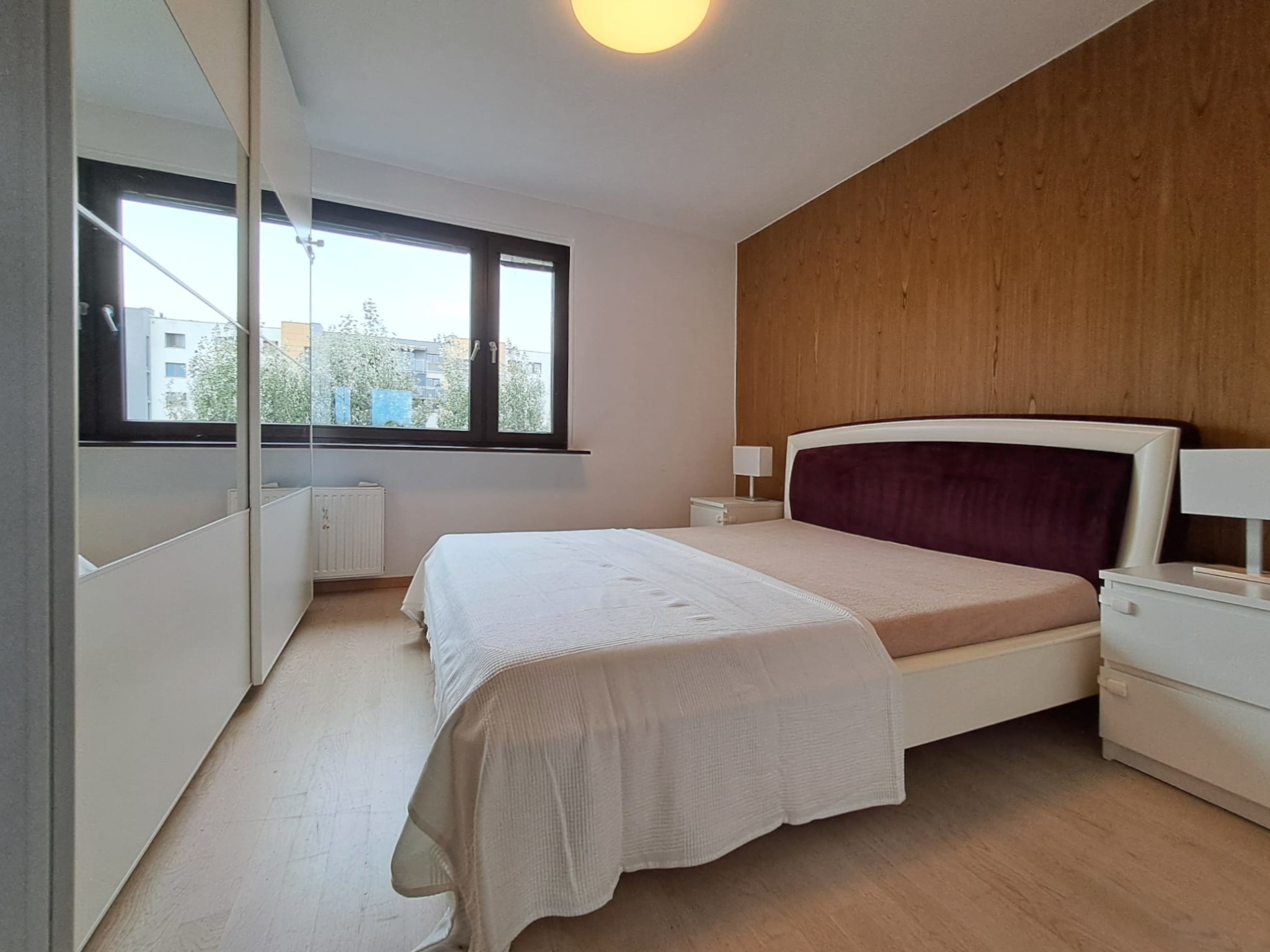 Jasna sypialnia z łóżkiem z bordowym zagłówkiem, drewnianą ścianą i oknem z widokiem na budynki.