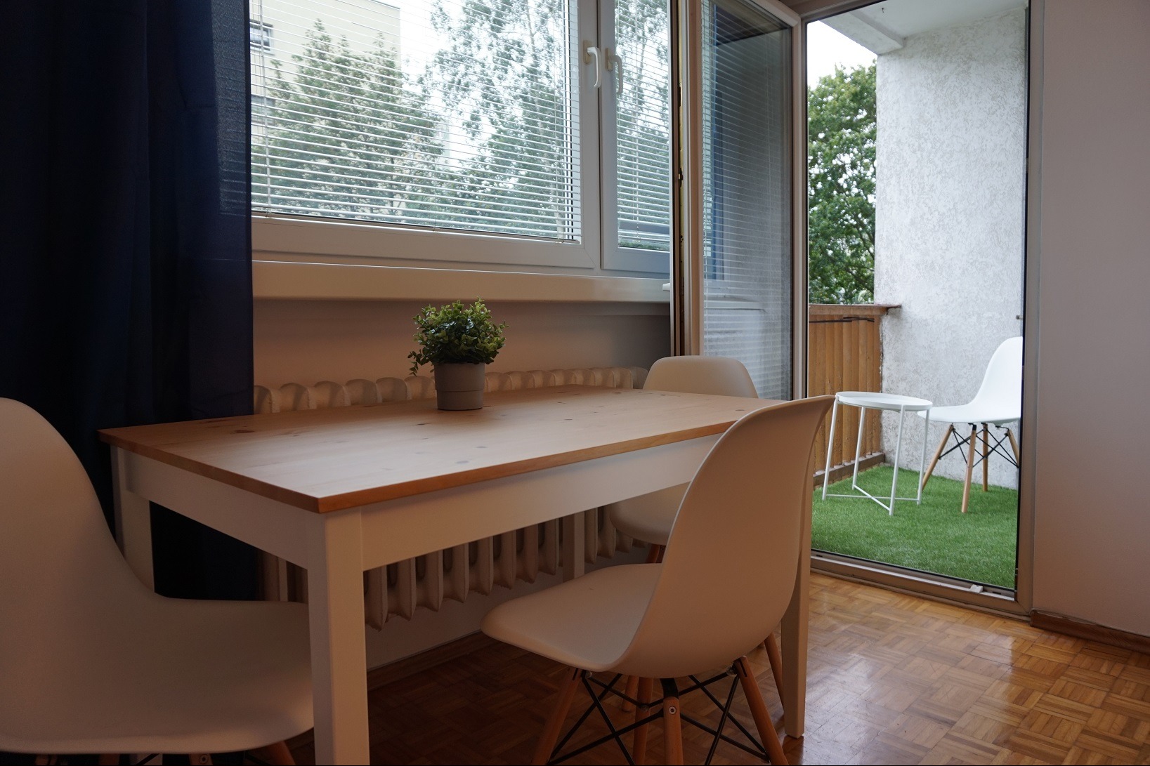 Jasne wnętrze z drewnianym stołem, trzema białymi krzesłami i rośliną doniczkową na tle okna z żaluzjami i balkonu z sztuczną trawą.