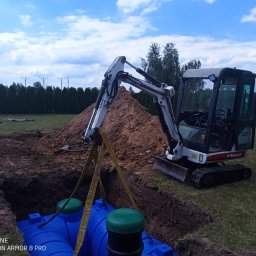 FU Bartosz Ludynia - Mini koparka Bobcat 322 opuszcza niebieski zbiornik z zielonymi pokrywami do wykopu, w tle pryzma ziemi, rów z ułożonymi pomarańczowymi rurami i zielony trawnik z tujami.