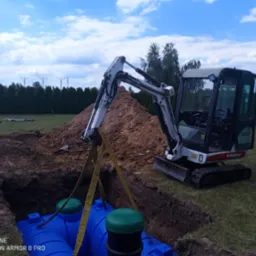 Mini koparka Bobcat 322 opuszcza niebieski zbiornik z zielonymi pokrywami do wykopu, w tle pryzma ziemi, rów z ułożonymi pomarańczowymi rurami i zielony trawnik z tujami.