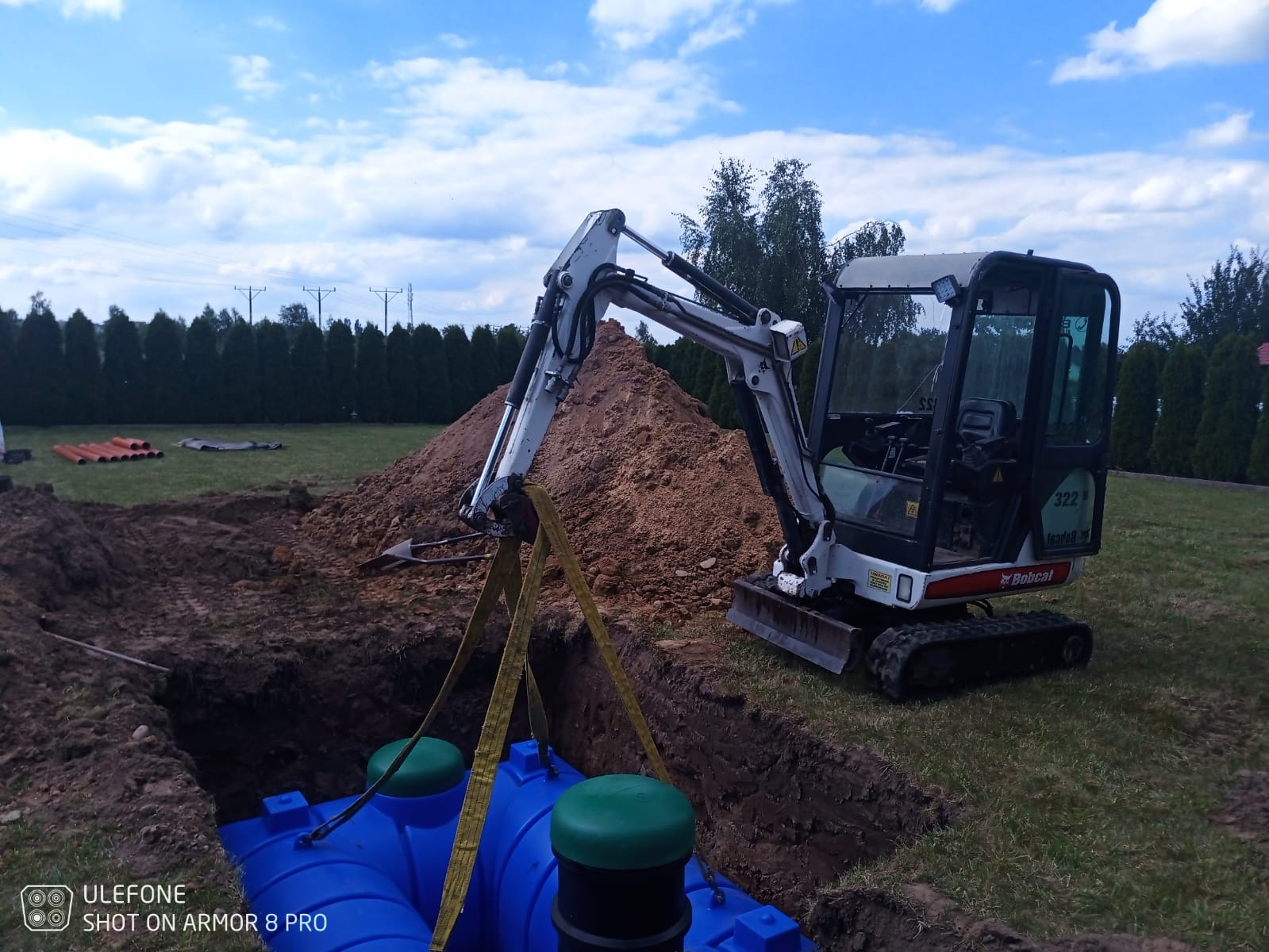 Mini koparka Bobcat 322 opuszcza niebieski zbiornik z zielonymi pokrywami do wykopu, w tle pryzma ziemi, rów z ułożonymi pomarańczowymi rurami i zielony trawnik z tujami.