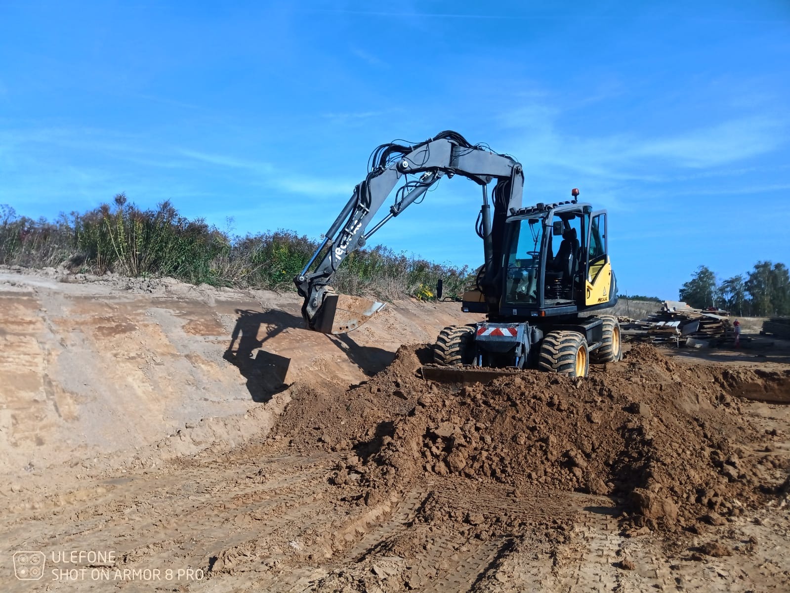 Żółto-szara koparka kołowa podczas prac ziemnych, widoczny wykop w gruncie i pryzma ziemi, błękitne niebo w tle.