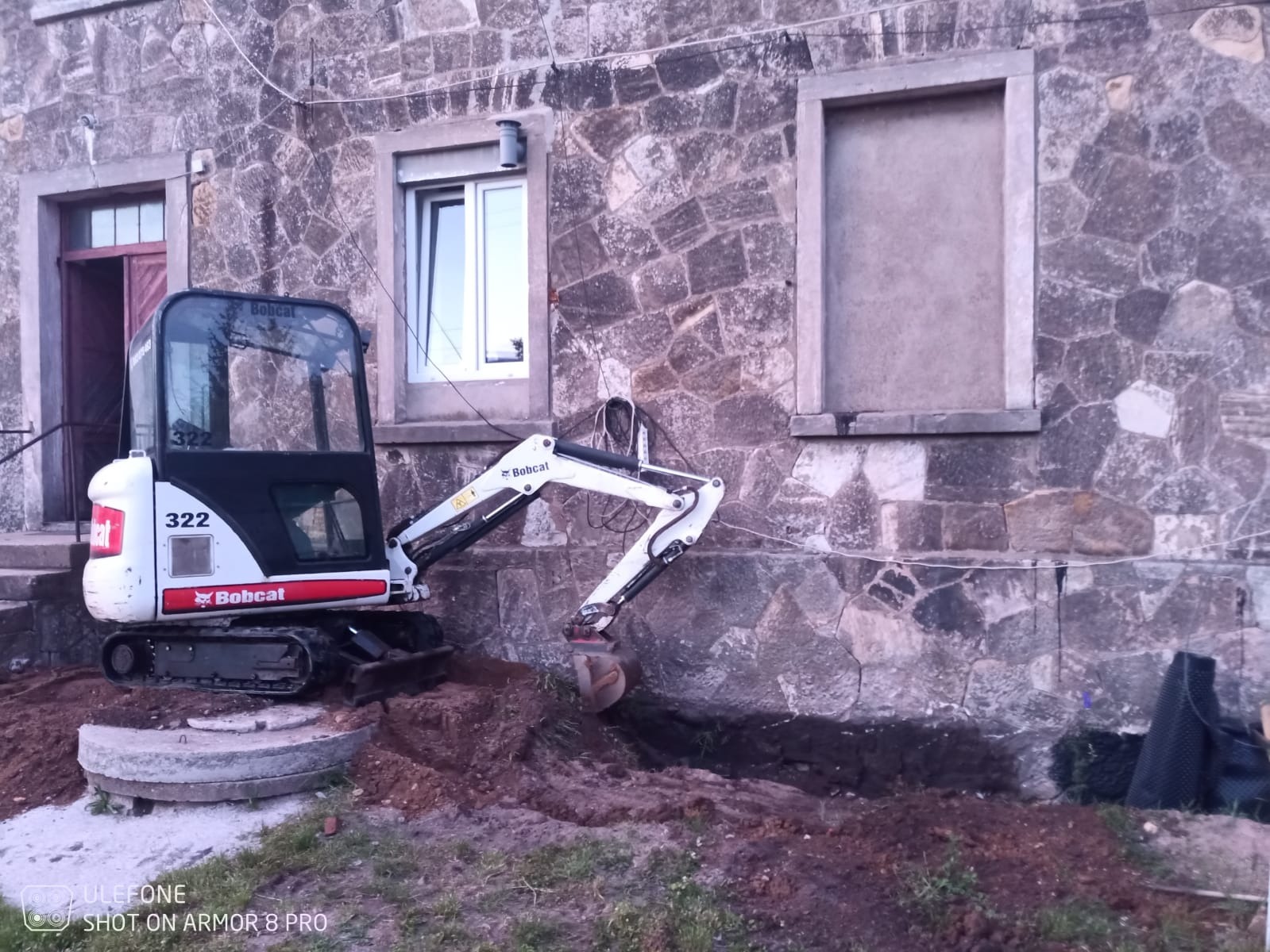 Mini koparka Bobcat 322 wykonuje wąski wykop wzdłuż kamiennej ściany budynku z oknami, widoczny betonowy właz kanalizacyjny