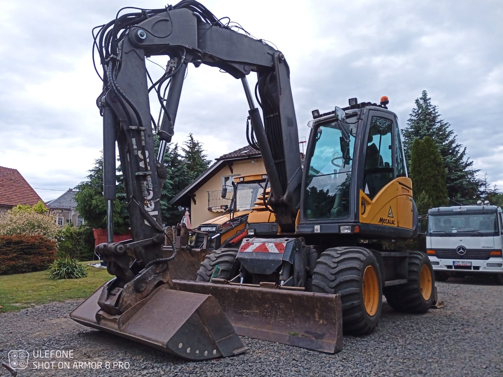Żółto-czarna koparka Mecalac stoi na żwirowym podłożu, z widocznym łyżką i ramieniem. W tle budynek, druga koparka Caterpillar i biała ciężarówka Mercedes.