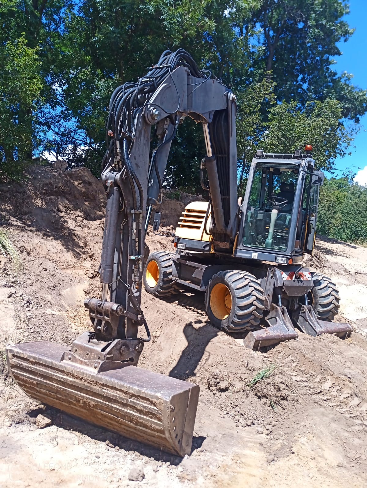 Kołowa koparka Volvo z łyżką na nierównym, piaszczystym terenie z widocznymi śladami pracy, w tle drzewa pod błękitnym niebem.