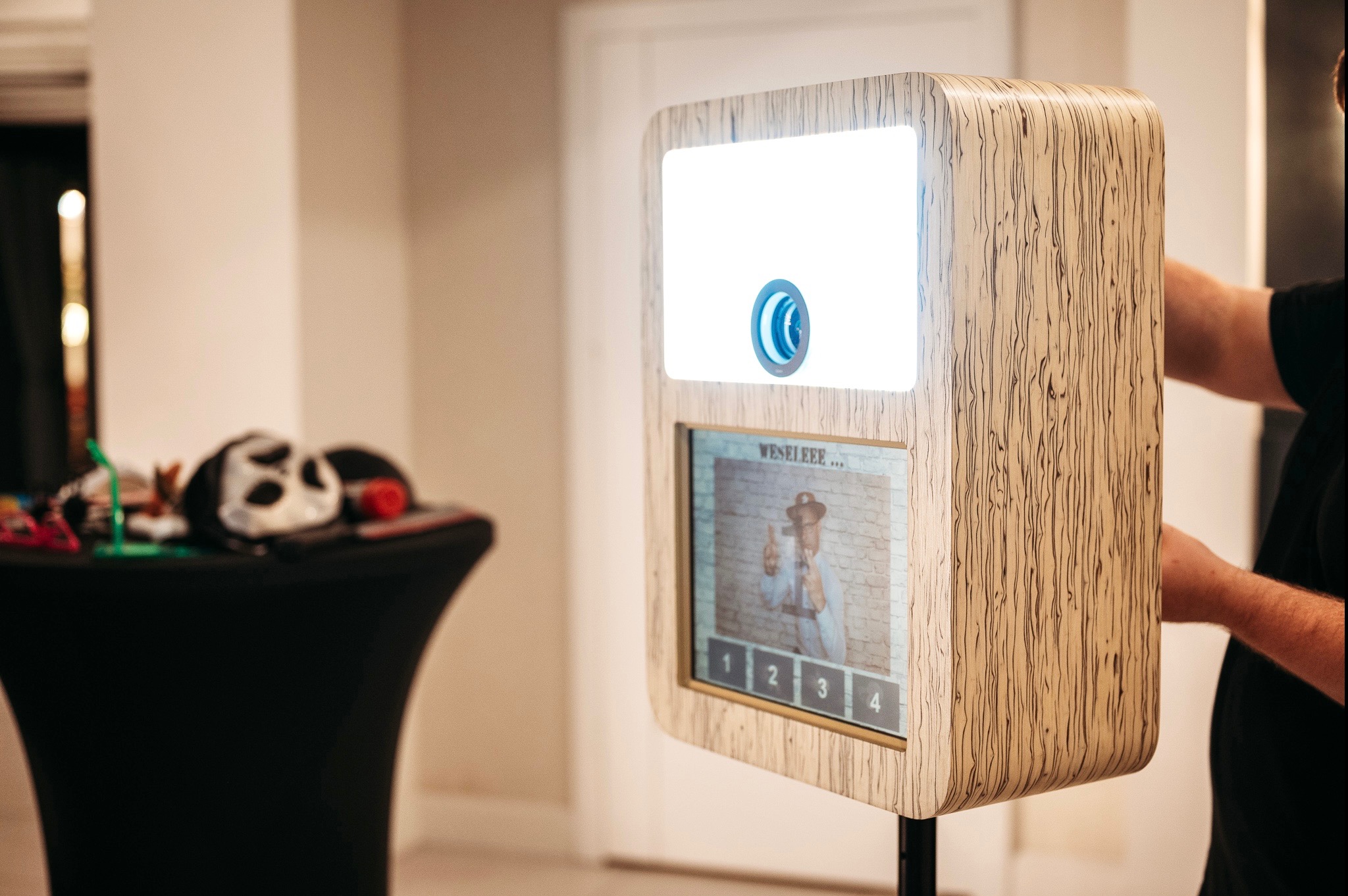 Drewniana obudowa automatu do zdjęć z ekranem dotykowym i rekwizytami na stole w tle.