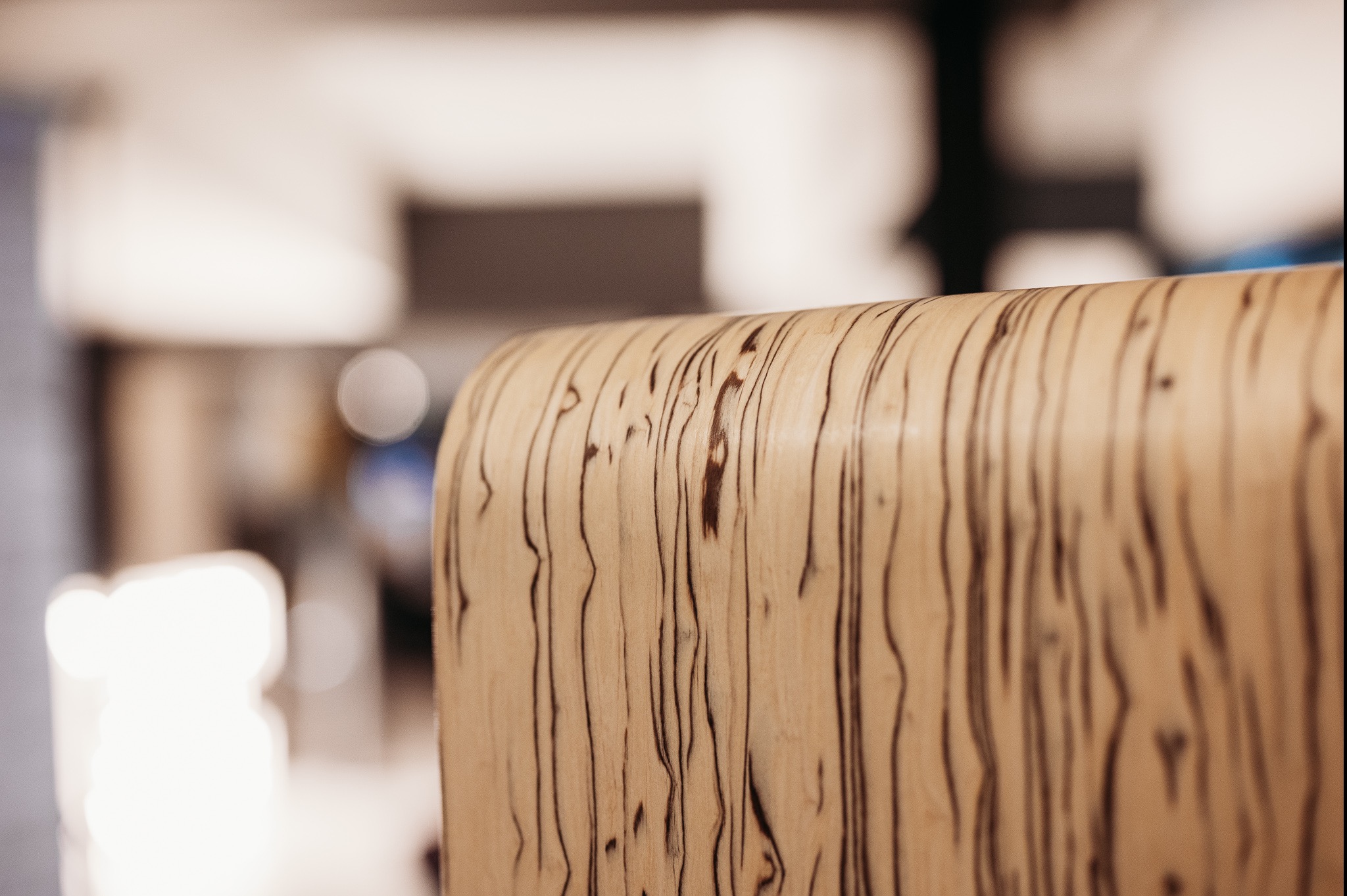 Fragment obudowy urządzenia z jasnego drewna z ciemnymi żyłkami na rozmytym tle wnętrza.