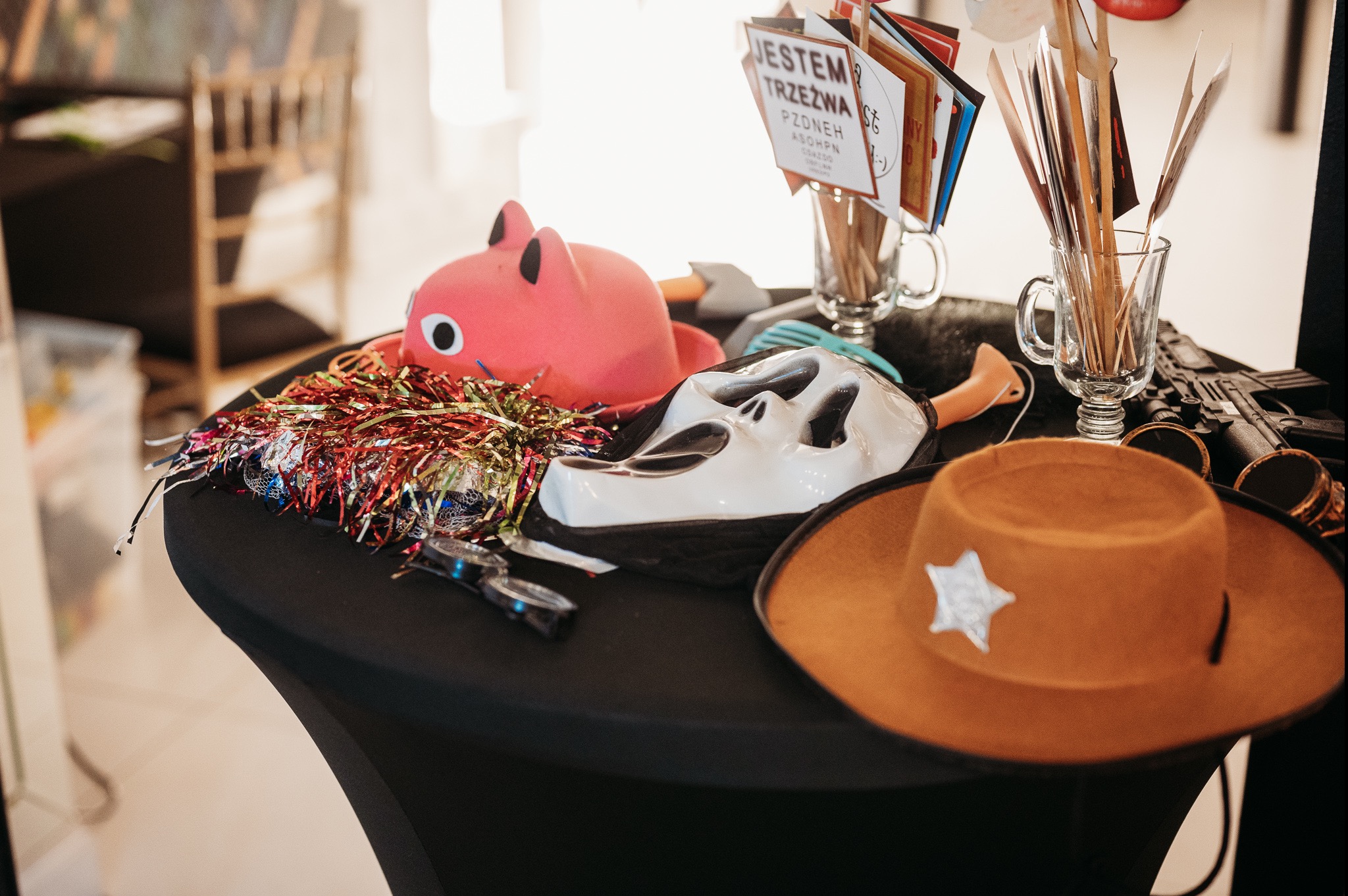 Akcesoria na czarnym stole: różowa maskotka świnki, biała maska, kowbojski kapelusz z gwiazdą szeryfa, okulary, serpentyna oraz rekwizyty do zdjęć w szklankach.