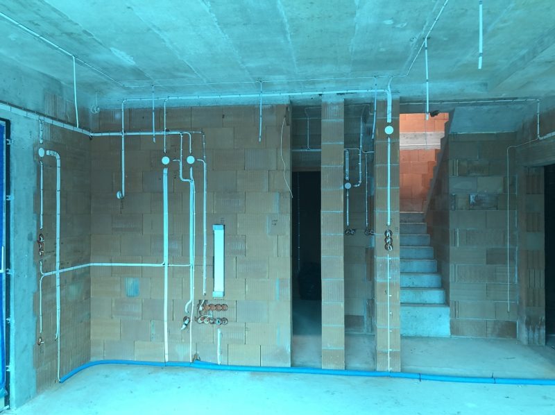 Surowy stan wnętrza z widocznymi instalacjami elektrycznymi poprowadzonymi po ścianach z bloczków, przewody w białych rurkach, niebieska rurka na podłodze, widoczne schody w tle.