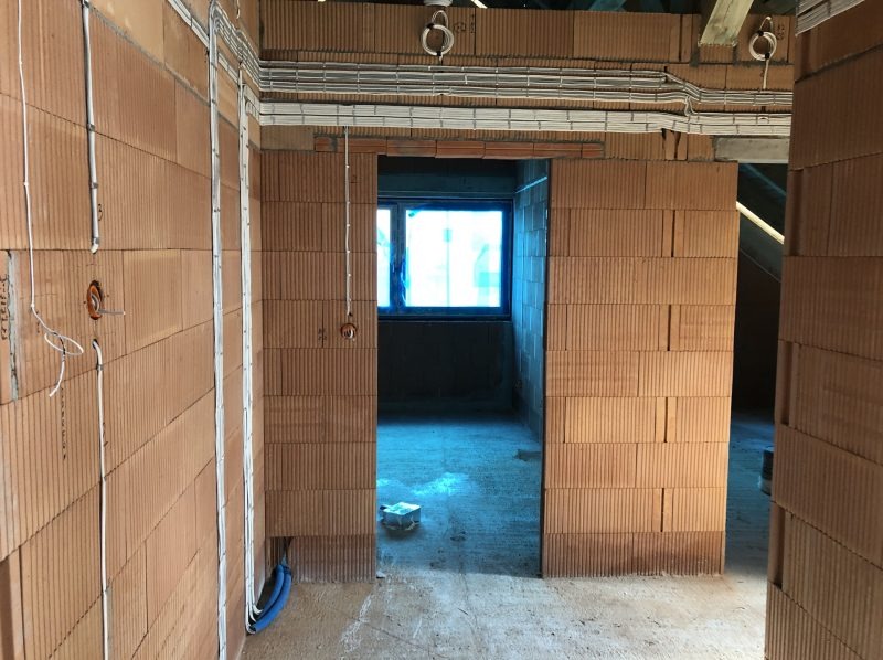 Surowy stan wnętrza z cegły ceramicznej z widocznymi przewodami elektrycznymi w korytkach na suficie i w ścianach, gniazdka elektryczne w ścianach, okno z niebieską folią ochronną.