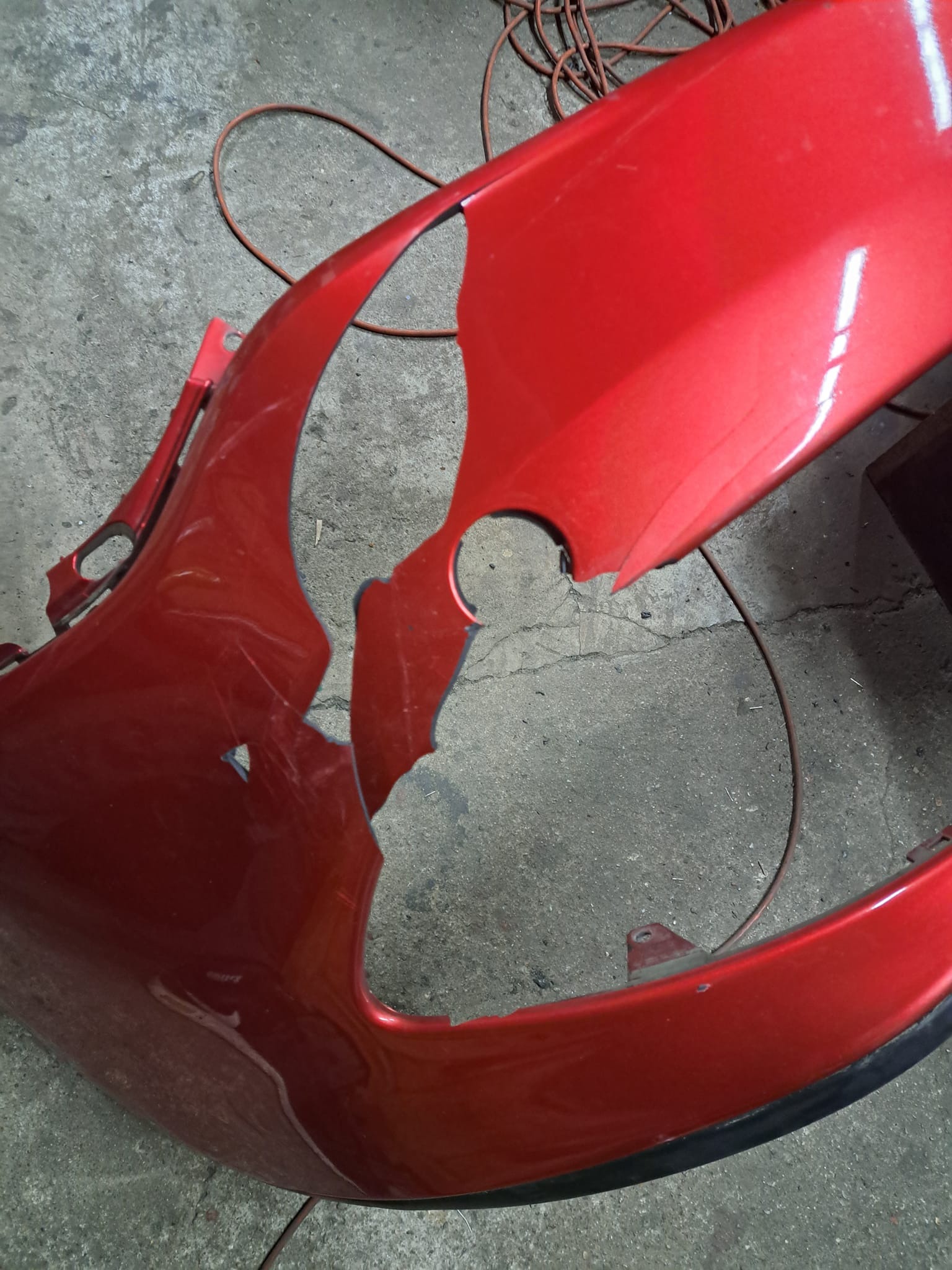 Uszkodzony, czerwony zderzak samochodowy z wyraźnymi pęknięciami i ubytkami, leżący na szarym betonowym podłożu z widocznym kablem w tle.