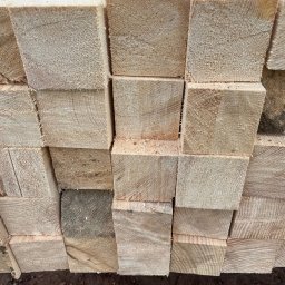 TARTAK Żywiec Wiezba Tarcica - Stos jasnych, oheblowanych belek z drewna budowlanego ułożonych w regularny sposób, widoczne ślady cięcia i drobne wióry.