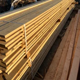 Stos jasnej, świeżo przetartej tarcicy budowlanej, zabezpieczony białą taśmą, z widocznymi śladami obróbki i drobnymi wiórami na powierzchni.