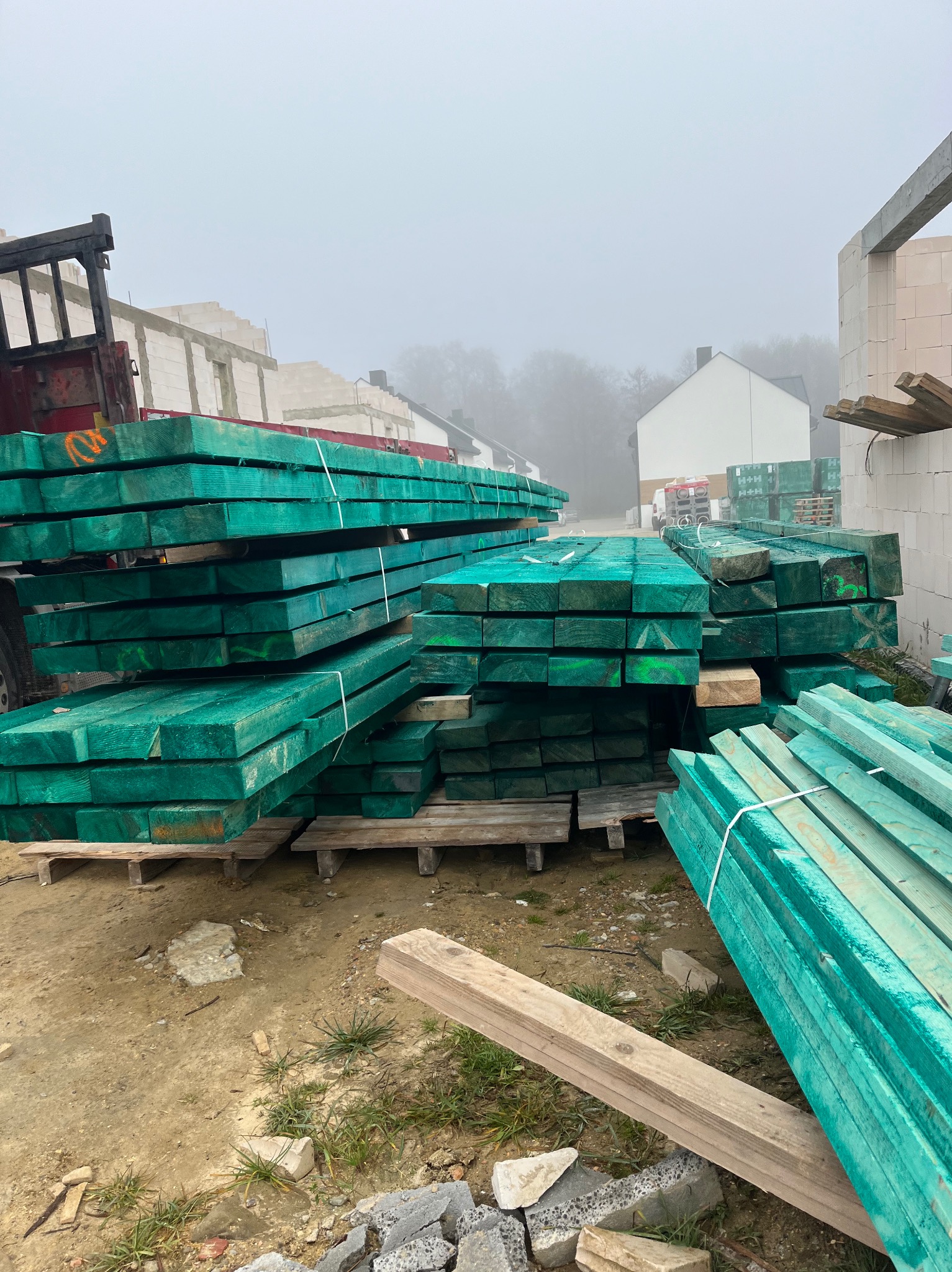 Stosy impregnowanej na zielono tarcicy budowlanej, zabezpieczonej białym sznurkiem, leżą na paletach na placu budowy w pochmurny dzień. W tle widoczny surowy budynek z białych bloczków i fragment...