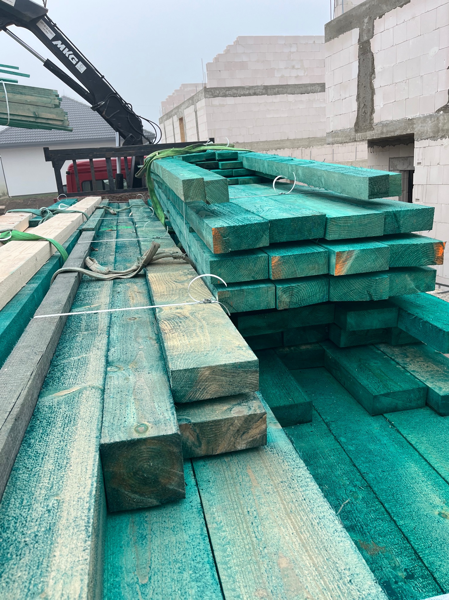 Świeżo zaimpregnowane na zielono drewniane belki ułożone na ciężarówce z dźwigiem, w tle budynek w stanie surowym.