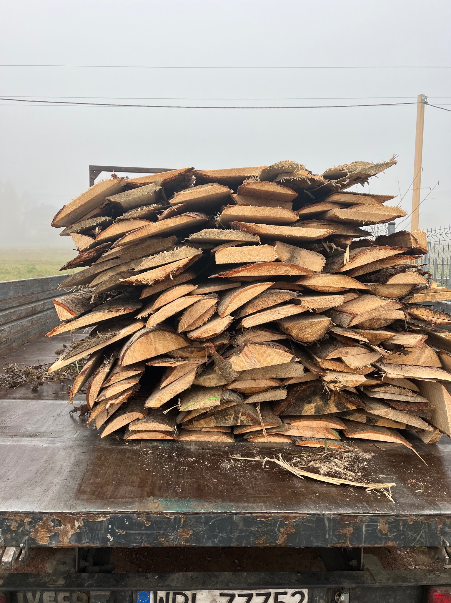 Stos nieociosanych desek ułożonych na pace samochodu, widoczny numer rejestracyjny pojazdu, w tle zamglony krajobraz.