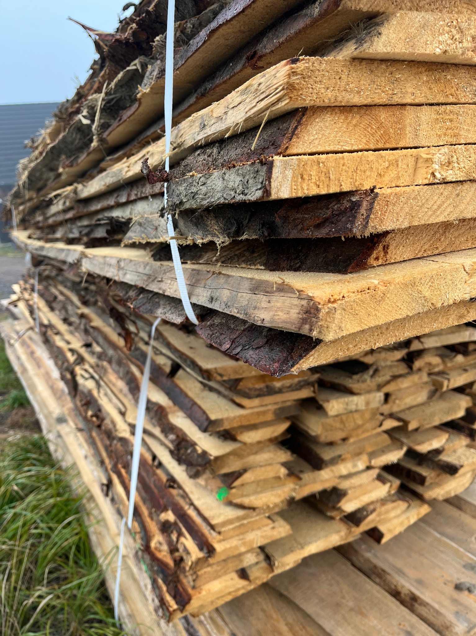 Stos świeżo przetartej tarcicy z widoczną korą, zabezpieczony białą taśmą, ułożony na trawie, z fragmentem budynku w tle.