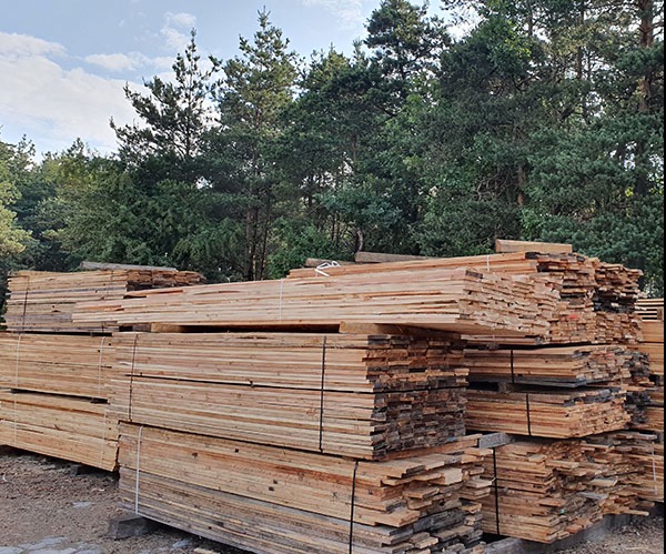 Stosy świeżo przetartej, jasnej deski na tle zielonego lasu, spięte białym sznurkiem.