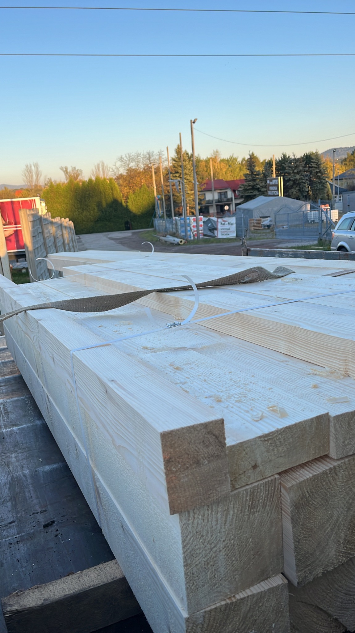 Świeżo przetarte, jasne, drewniane belki budowlane, zabezpieczone pasami transportowymi, leżące na ciemnej powierzchni transportowej, z widocznym fragmentem zabudowań w tle.