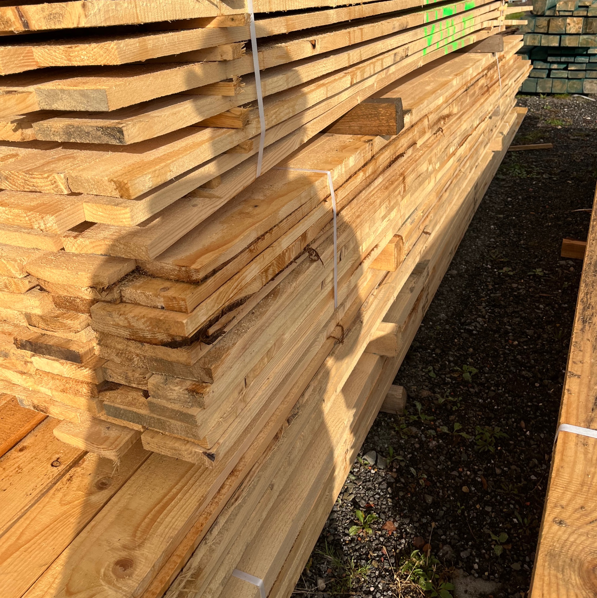 Stos jasnej, świeżej tarcicy budowlanej, związanej białymi taśmami, ułożony na żwirowym podłożu z zielonymi roślinami, z widocznymi zielonymi oznaczeniami na górnej warstwie.