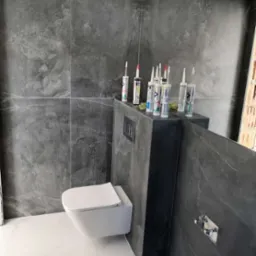 Nowoczesna łazienka z szarymi płytkami imitującymi kamień i podwieszaną toaletą. Na ściance obudowującej stelaż widoczne tuby z silikonem i włącznik światła.