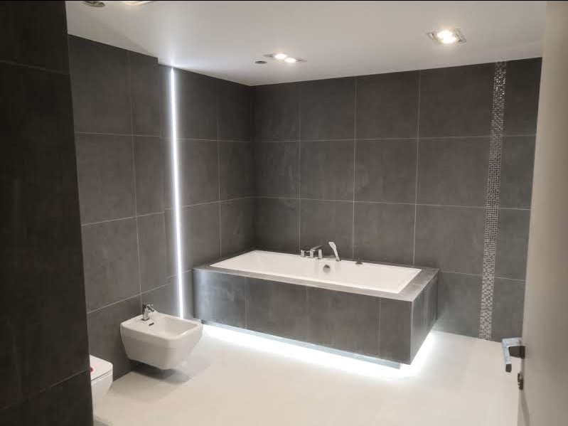 Nowoczesna łazienka z szarymi płytkami, podświetloną wanną i pionową listwą LED, minimalistyczny design.