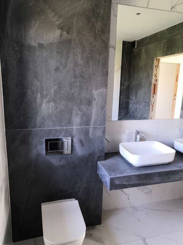 Wykończona łazienka z szarymi płytkami imitującymi marmur na ścianach, podwieszaną toaletą z chromowanym przyciskiem spłukiwania oraz umywalką nablatową na blacie z tego samego materiału co ściany.