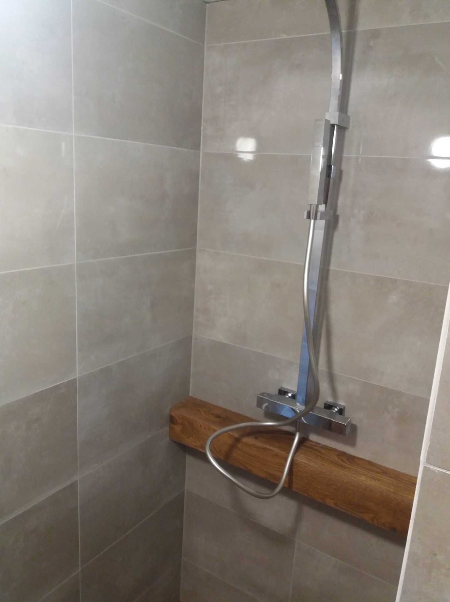 Nowoczesny prysznic z drewnianą półką w łazience wyłożonej szarymi płytkami.