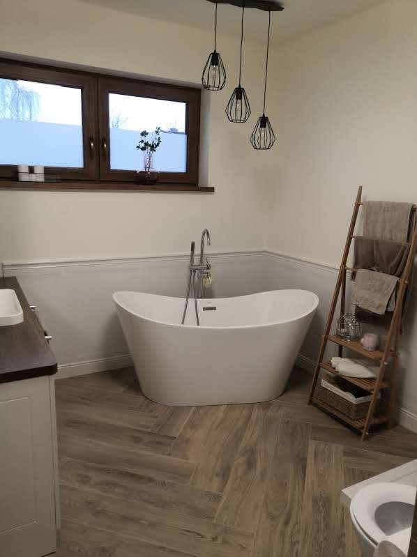 Nowoczesna łazienka z wolnostojącą białą wanną, drewnopodobną podłogą ułożoną w jodełkę, drewnianą drabinką na ręczniki i designerskimi lampami wiszącymi.