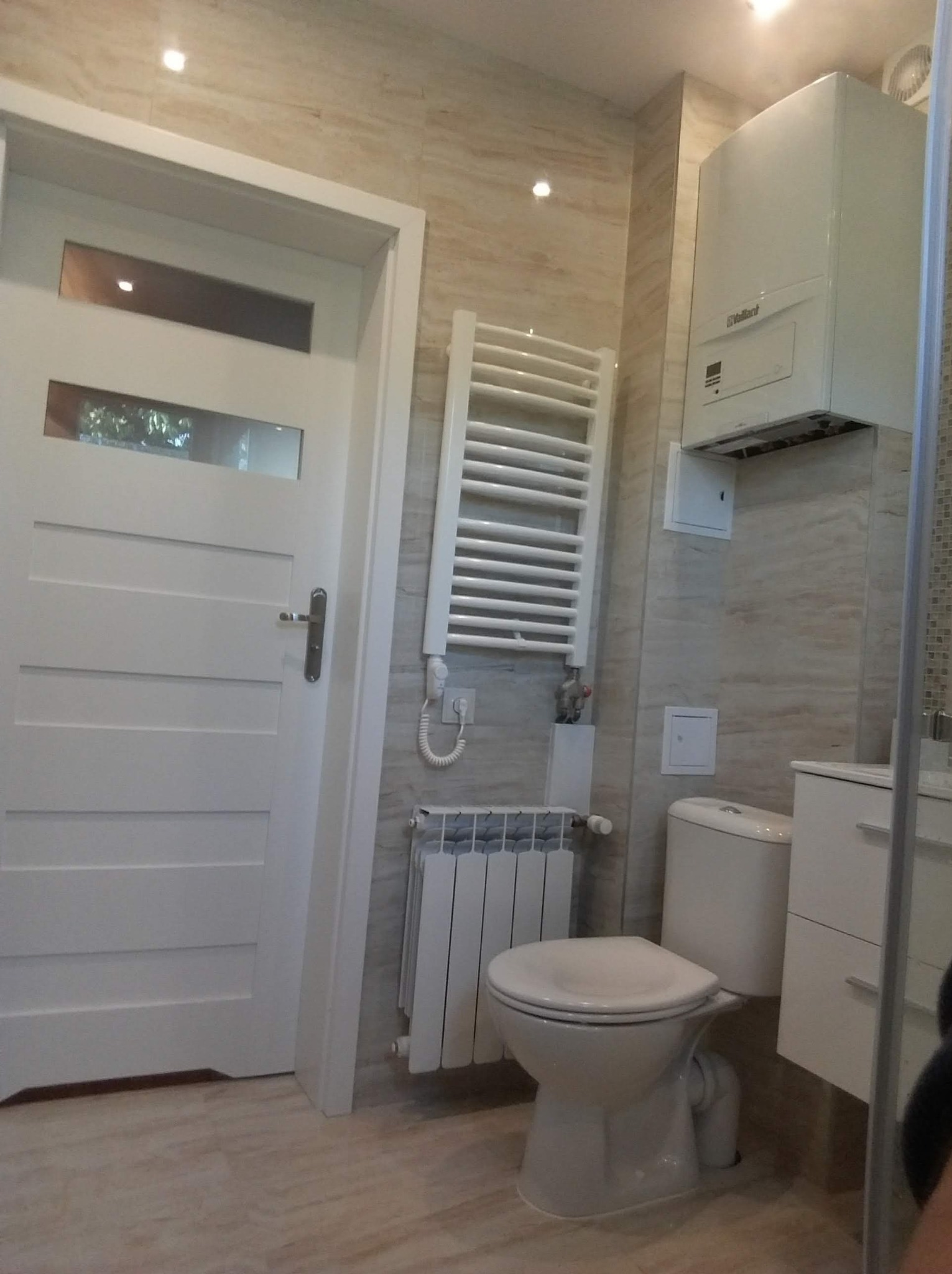 Biała toaleta kompaktowa, grzejnik drabinkowy i piec gazowy w wykończonej łazience z beżowymi płytkami i białą stolarką drzwiową.