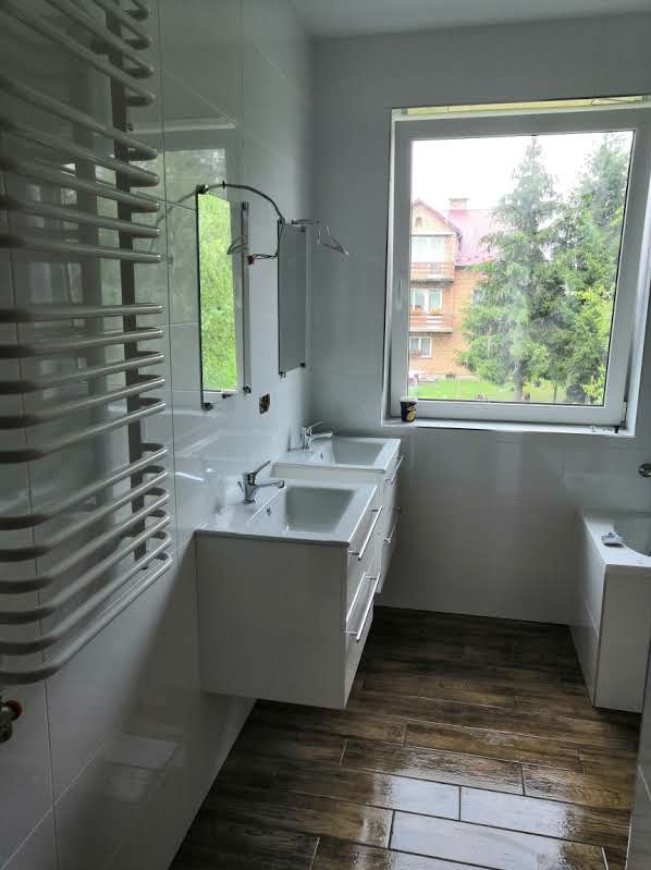 Jasna łazienka z dwoma umywalkami na szafkach z szufladami, grzejnik drabinkowy na ścianie, okno z widokiem na budynek i drzewa, podłoga wyłożona drewnopodobnymi płytkami.
