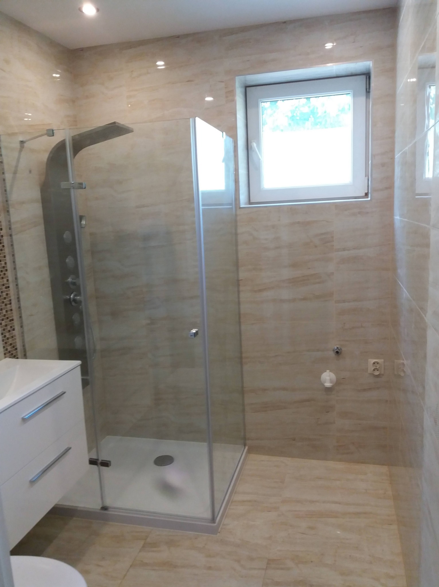 Nowoczesna kabina prysznicowa z panelem natryskowym w łazience wykończonej beżowymi płytkami imitującymi marmur, z oknem z widokiem na zieleń.