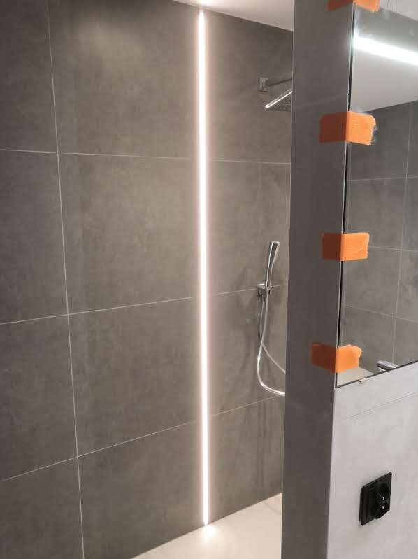 Nowoczesna łazienka z szarymi płytkami, pionowym oświetleniem LED w ścianie i szklaną kabiną prysznicową zabezpieczoną taśmą malarską.