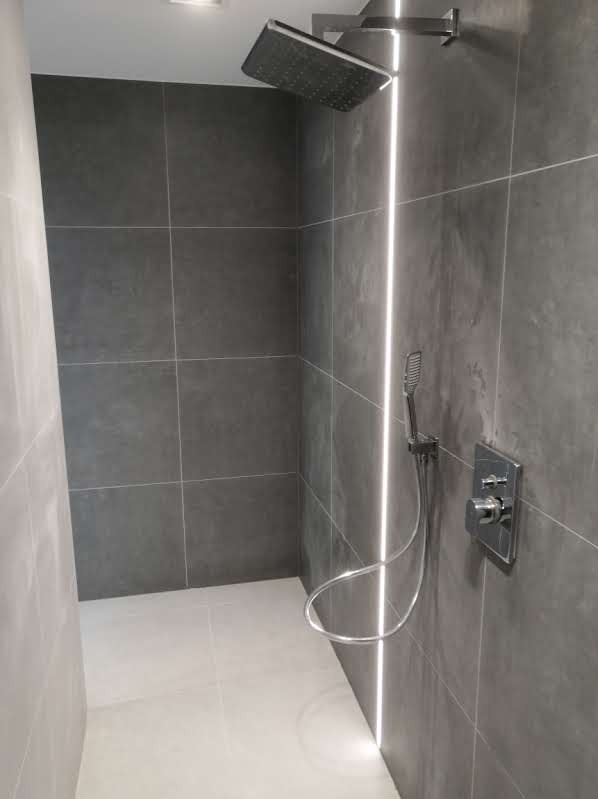 Nowoczesna kabina prysznicowa z szarymi, kwadratowymi płytkami, oświetleniem LED w pionowej szczelinie i chromowaną armaturą. Widoczny deszczownica, słuchawka prysznicowa oraz bateria termostatyczna.