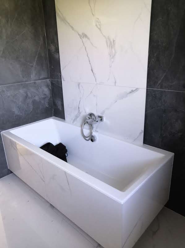 Nowoczesna, prostokątna wanna obłożona płytkami imitującymi marmur, z chromowaną baterią i czarnym materiałem wewnątrz, na tle ściany wykończonej szarymi i białymi płytkami.