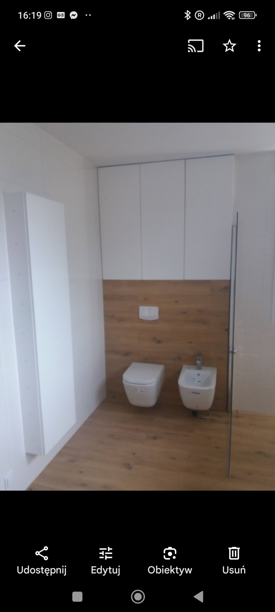 Nowoczesna łazienka z wiszącą toaletą i bidetem na tle drewnopodobnej ściany, białe szafki wiszące i podłoga w tym samym odcieniu drewna.