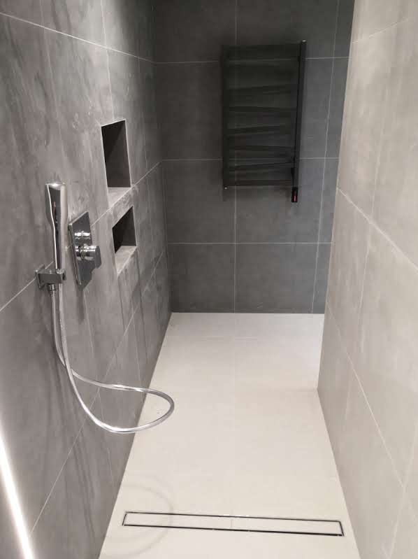 Nowoczesna łazienka z szarymi płytkami, wnękami w ścianie, chromowaną armaturą i odpływem liniowym w białej posadzce bez brodzika.