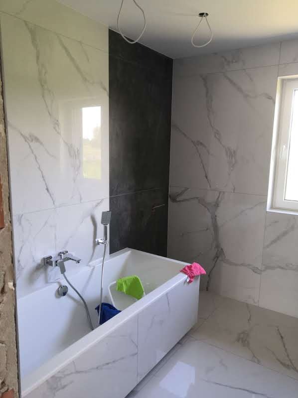 Wanna obudowana płytkami imitującymi marmur, z widoczną armaturą i fragmentem ściany w ciemnym kolorze. W tle surowe przewody elektryczne i okno.