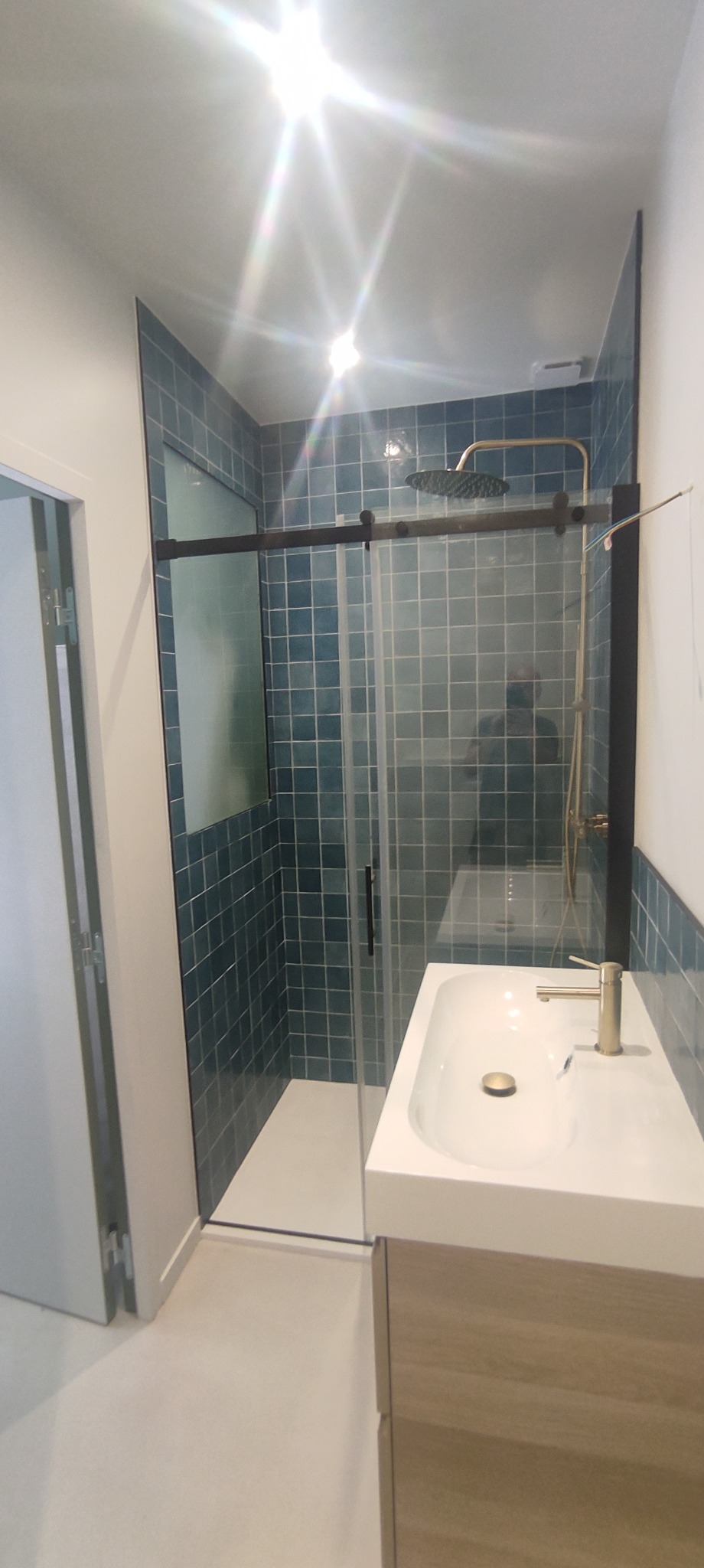 Nowoczesna łazienka z kabiną prysznicową typu walk-in, wykończona niebieską mozaiką i złotą armaturą, umywalka nablatowa na szafce z drewnianym frontem.