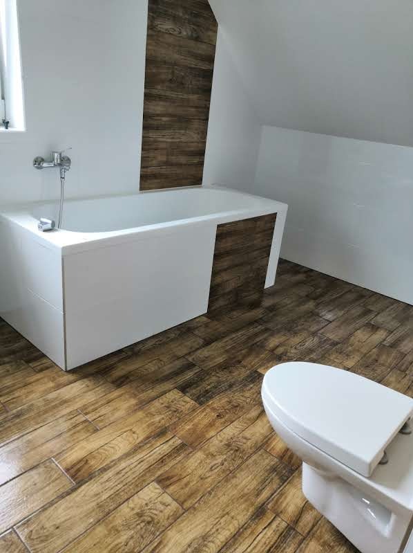 Biała wanna z baterią, toaleta i imitująca drewno okładzina na podłodze i części ściany w łazience na poddaszu.