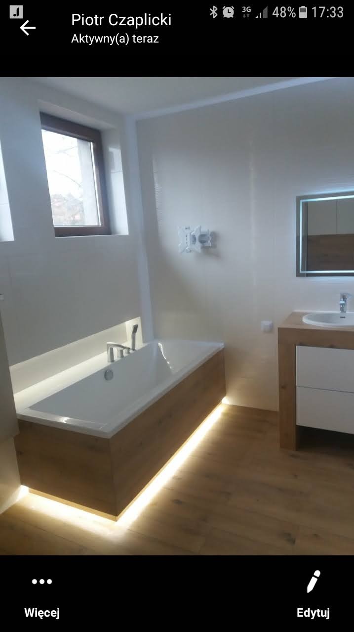 Nowoczesna łazienka z podświetlaną wanną obudowaną drewnem, drewnianą szafką z umywalką i lustrem z podświetleniem LED, wykończona w jasnych kolorach.