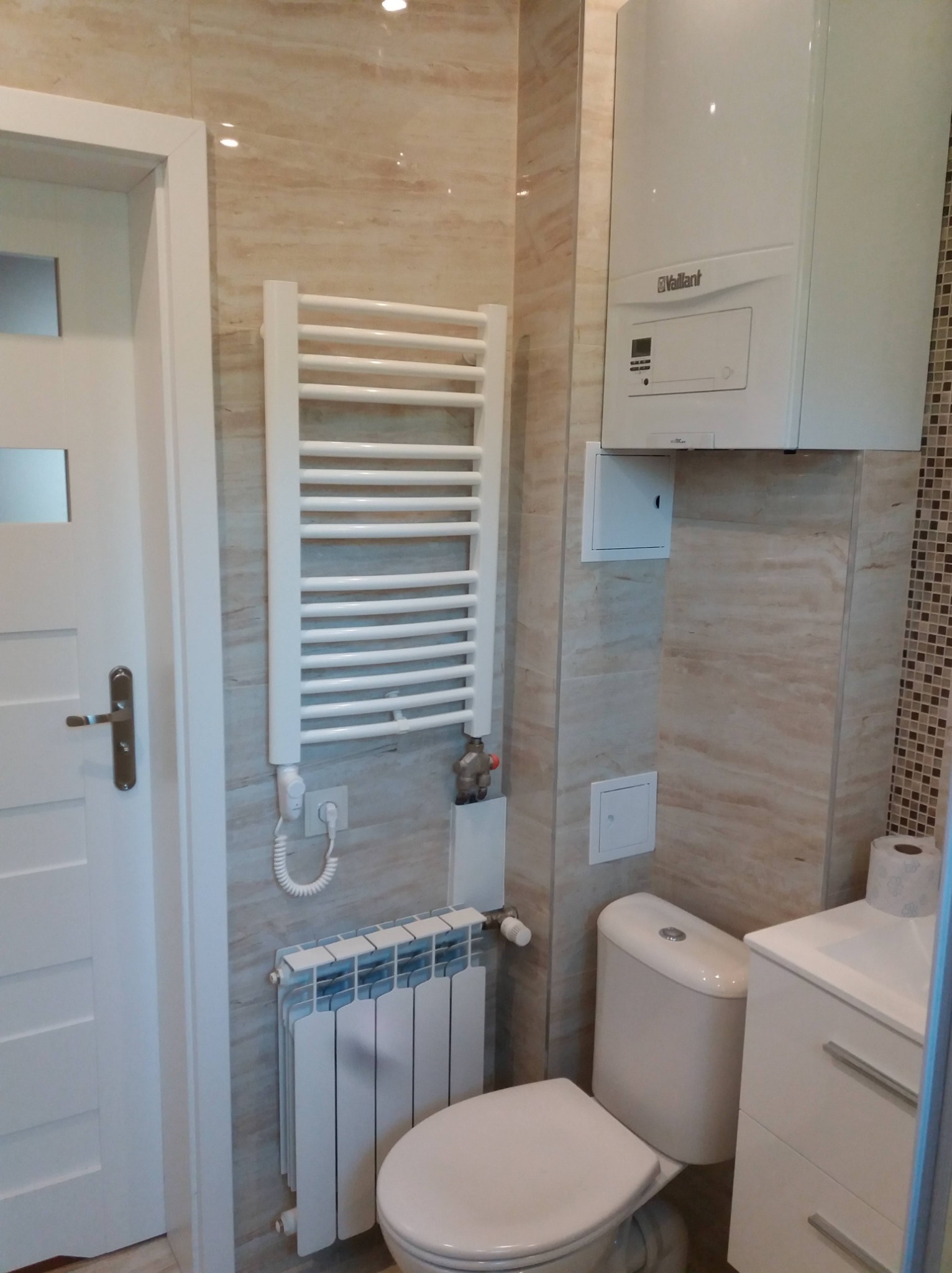Wykończona łazienka z beżowymi płytkami imitującymi marmur, białą armaturą, grzejnikiem drabinkowym, bojlerem Vaillant i grzejnikiem żeberkowym pod nim.